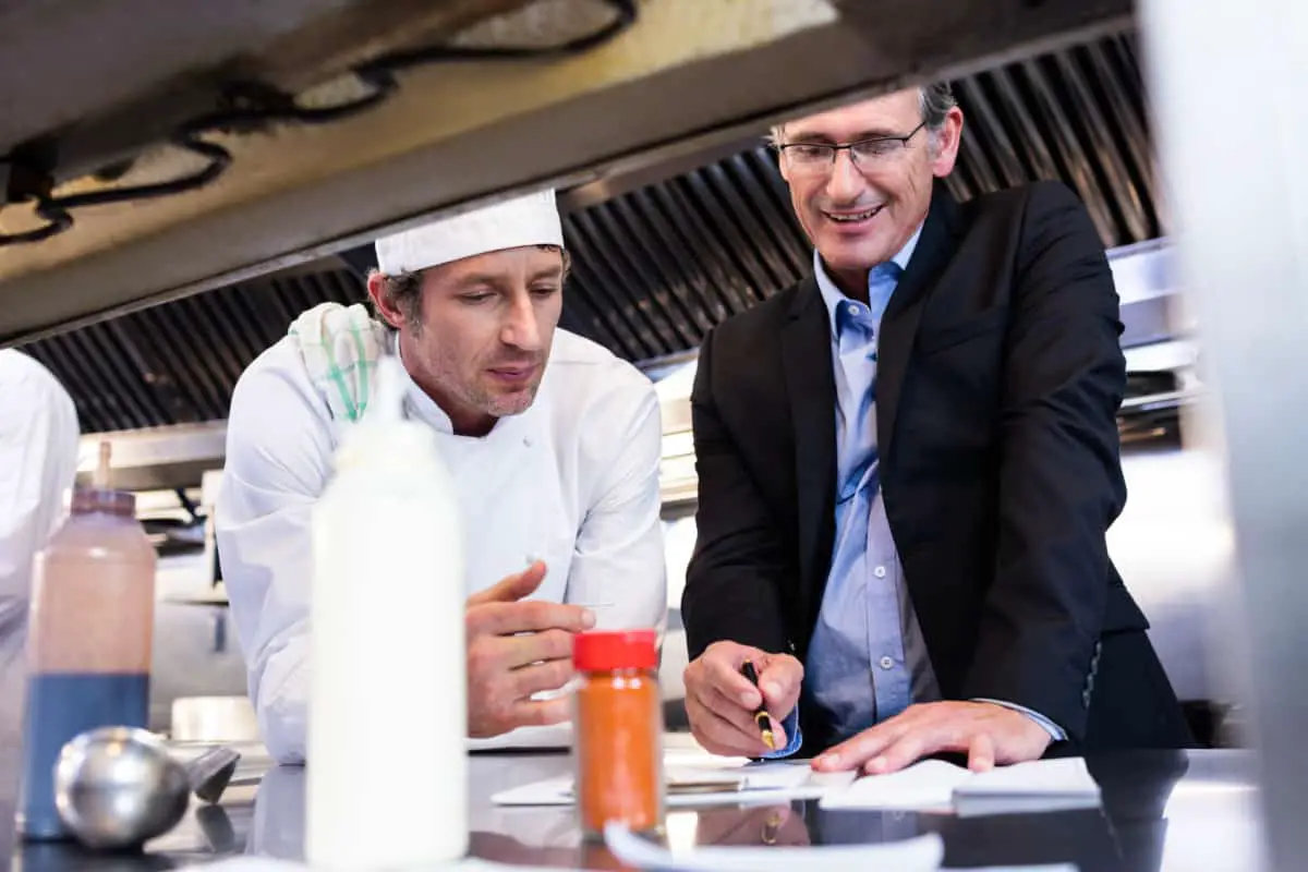 Canva Male Restaurant Manager Writing On Clipboard While Interacting To Head Chef 